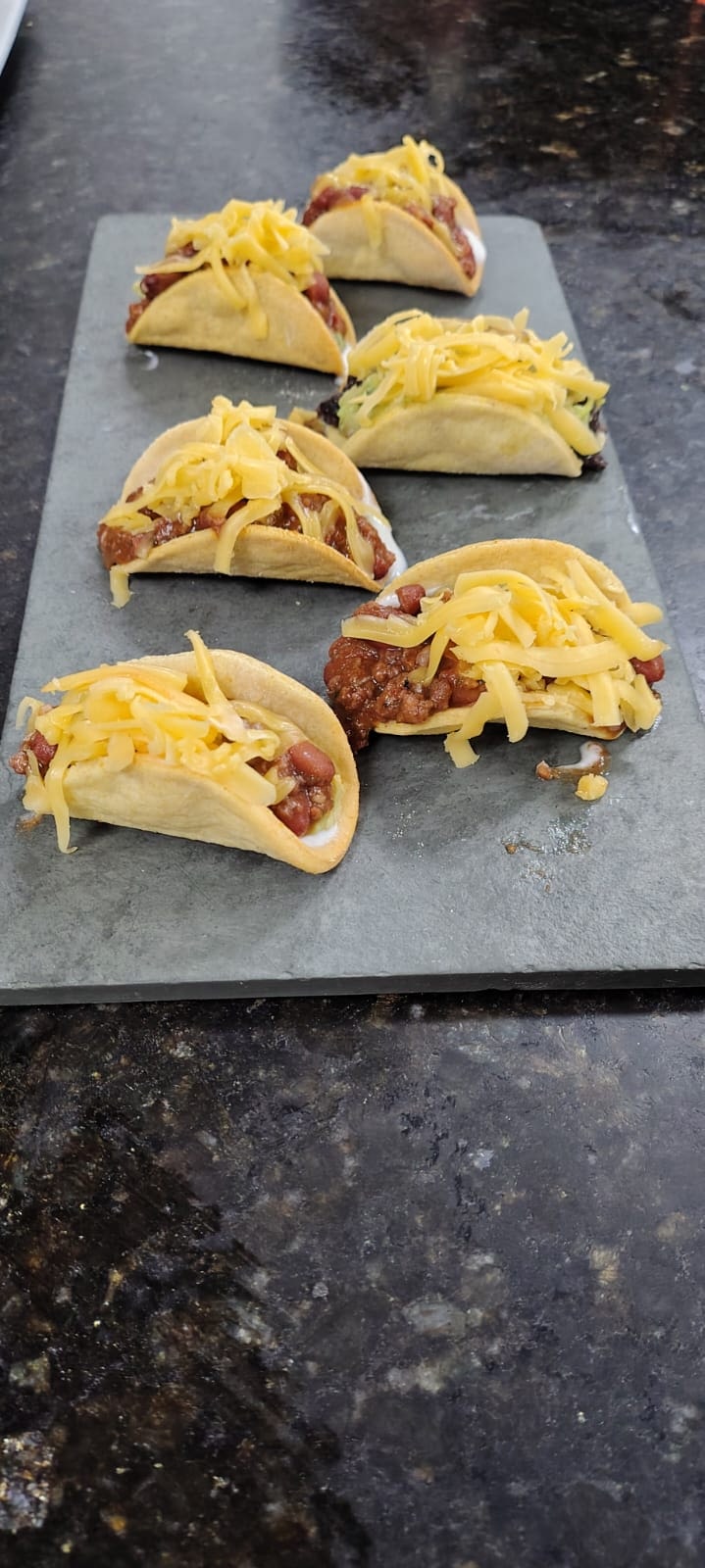 Seis tacos recheados e finalizados com queijo sobre uma tábua escura.